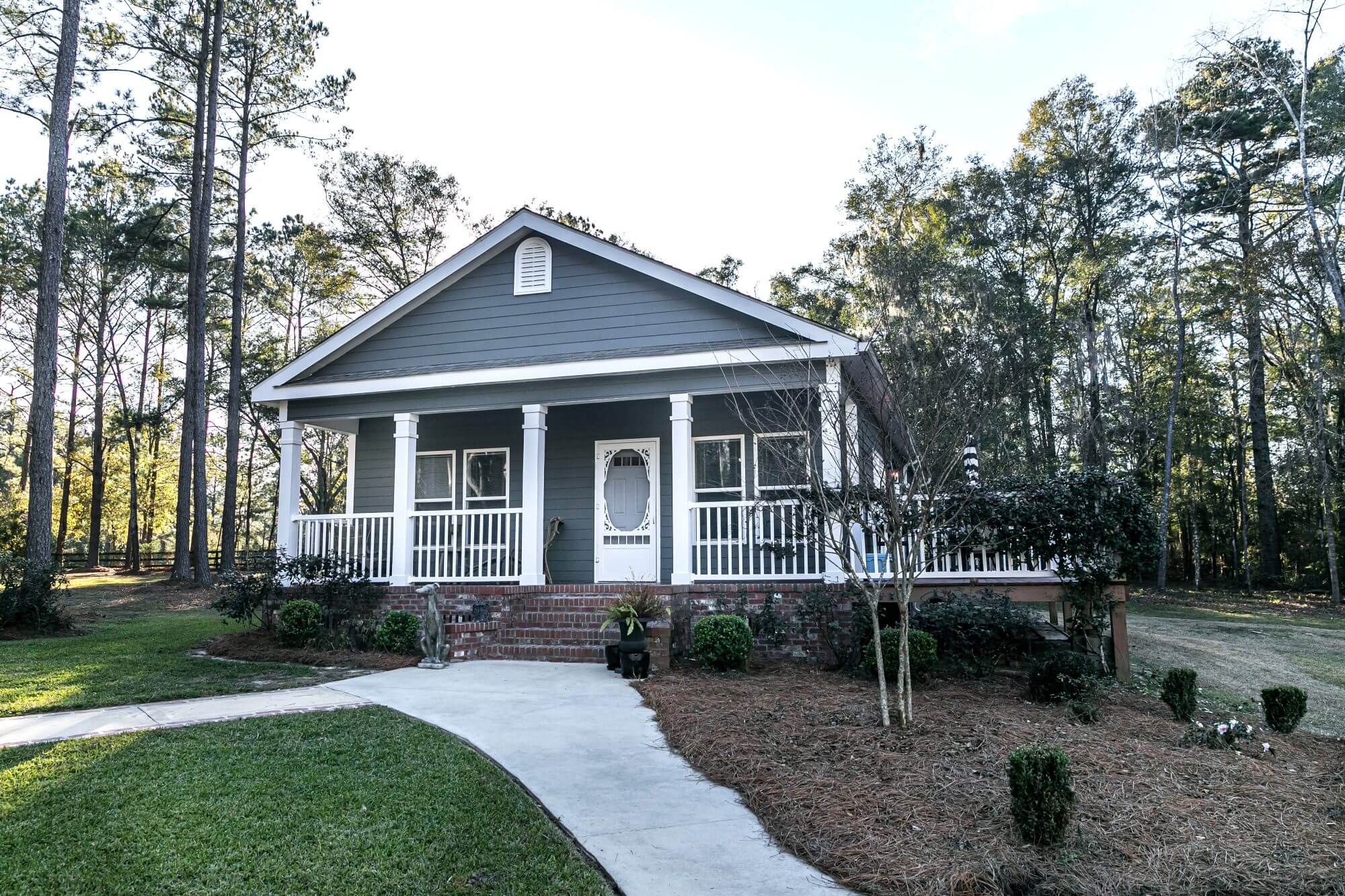 Small blue gray mobile home with a front and side porch , white railing out in the country on land shared by a larger siding house.