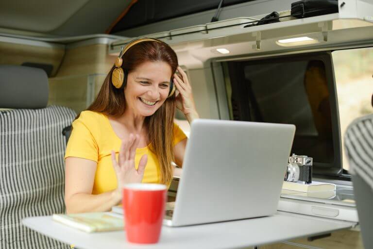 Cheerful female traveler in headphones waving hand and having video chat via laptop while sitting at table in camper