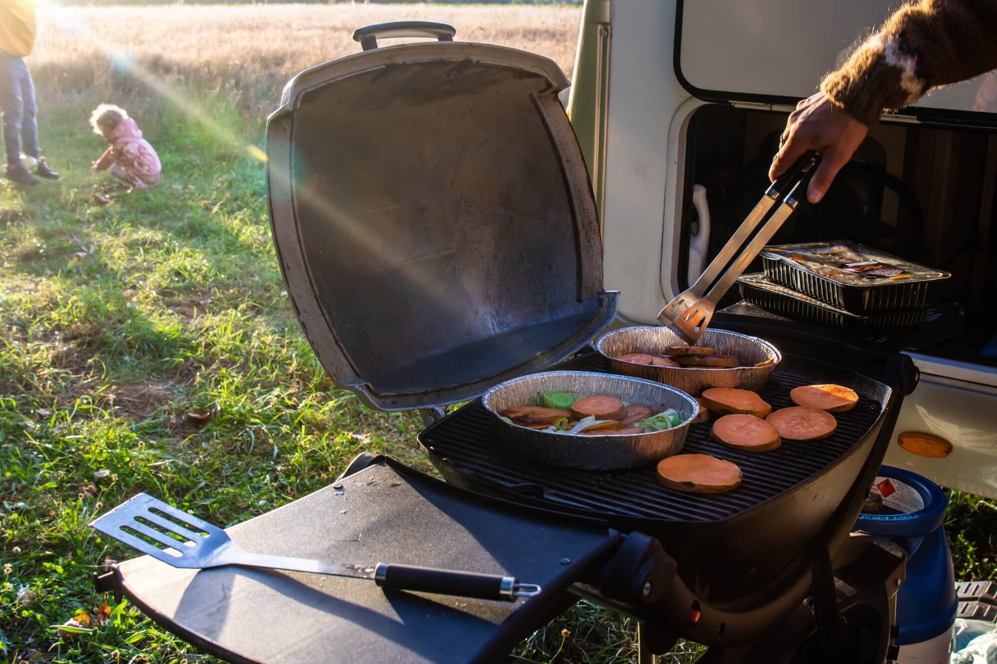 A hand with barbecue tongs grilling food on a gas grill. Kids playing in the background. Sun is shining. Part of mobile home in the background.