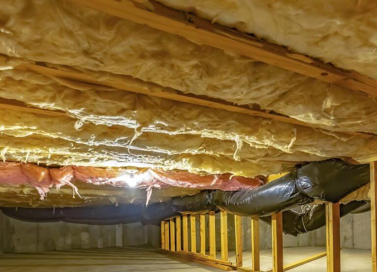 Basement or crawl space with upper floor insulation and wooden support beams. An area of limited height under the floor of a house with concrete wall and floor.