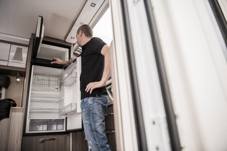 Broken RV Camper Travel Trailer Refrigerator. Caucasian Technician Trying to Fix the Appliance Inside Modern Recreational Vehicle.